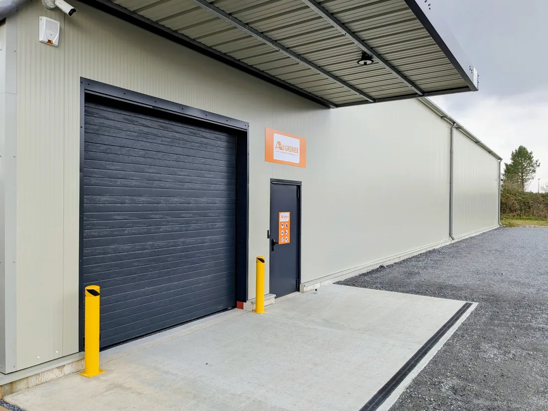 L'entrée du bâtiment Au Grenier au Barp : porte sectionnelle, auvent pour décharger à l'abri et enseigne orange